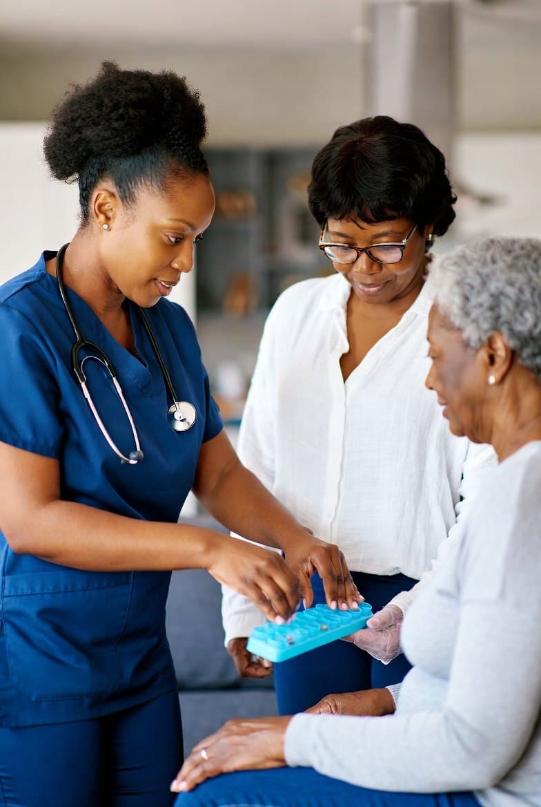 A nurse and a daughter help an older woman fill her pill organiser